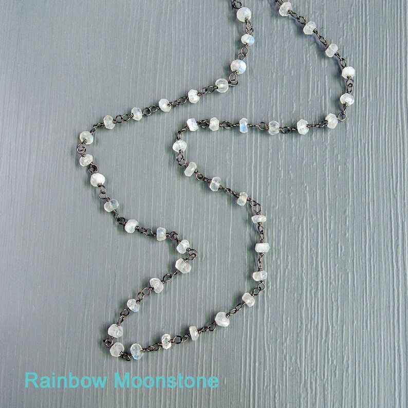 May include: A silver chain necklace with rainbow moonstone beads. The necklace is on a light gray wooden surface.