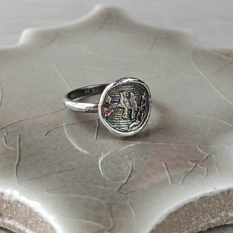 May include: A silver ring with a round, textured top featuring a detailed design of two birds perched on a branch. The ring is on a light gray, textured surface.