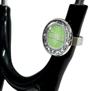 Puede incluir: Un charm de estetoscopio plateado con un diseño verde y negro. El charm tiene el nombre "Tarrah" escrito en él.