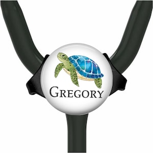 May include: A black stethoscope ID tag featuring a circular white insert with a watercolor illustration of a green and blue sea turtle. The name GREGORY is printed in black serif capital letters below the turtle on the white background.