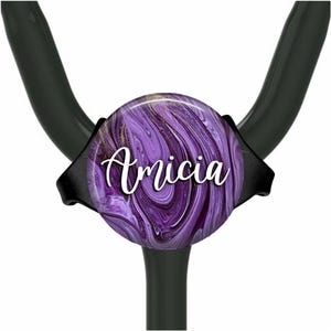 Puede incluir: Encanto de estetoscopio de mármol morado y dorado con el nombre "Amicia" escrito en blanco.