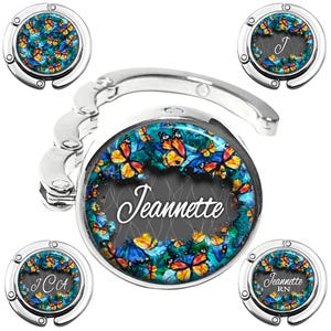 May include: A silver-toned metal foldable purse hook featuring a circular insert with a vibrant butterfly pattern in shades of blue, orange, and yellow. The center displays the name Jeannette in white script, with variations showing J, JCA, and Jeannette RN.