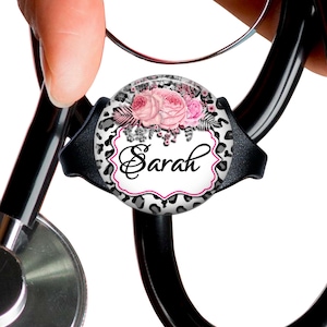 May include: A black stethoscope with a round, white and black patterned charm. The charm features a pink floral design and the name "Sarah" in black lettering.