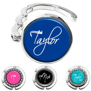 May include: A silver-toned foldable purse hook featuring a circular center with a deep blue background and the white script name Taylor. Three additional smaller purse hooks are shown below in pink, black, and turquoise with custom text.