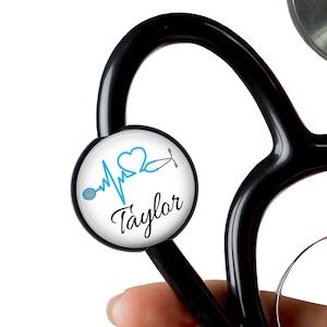 Puede incluir: Estetoscopio negro con un charm redondo blanco adjunto. El charm tiene una línea de latido del corazón azul, un icono de estetoscopio y el nombre "Taylor" escrito en negro.