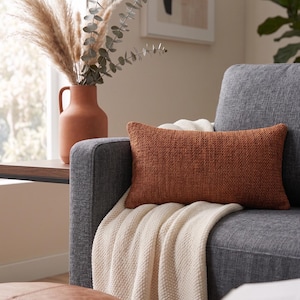 May include: A rectangular rust-colored pillow with a textured weave sits on a gray sofa, draped with a cream-colored knitted throw. A terracotta vase holds dried plants, and a leather ottoman is in the foreground.