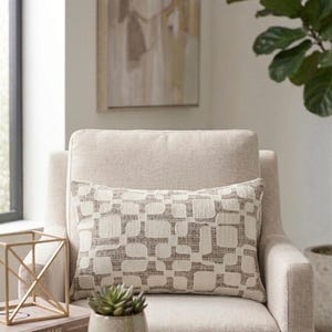 May include: A beige armchair with a rectangular pillow featuring a geometric pattern in shades of beige and brown. A small succulent in a ceramic pot sits on a wooden side table. A gold geometric sculpture and books are also on the table.