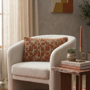 May include: A cozy living room scene featuring a cream-colored armchair with a textured, rust-colored pillow. A small wooden side table holds books, a succulent, and a candle. The room has a warm, inviting aesthetic with neutral tones.
