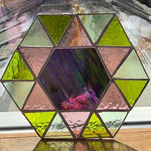 Puede incluir: Un atrapasoles geométrico de vidrieras con forma hexagonal. El diseño presenta triángulos en tonos verdes, rosas y una sección central con tonos morados y rosas. Se adjunta una cinta dorada para colgarlo.