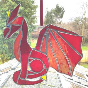 May include: A stained glass dragon ornament in shades of red, orange, and yellow. The dragon has a geometric design with a red ribbon for hanging. The dragon's wings are spread, and it has a yellow accent on its foot.