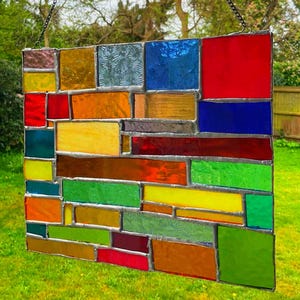 Puede incluir: Un colorido panel de vidrieras con una variedad de formas rectangulares y cuadradas en tonos de rojo, naranja, amarillo, verde y azul. Las piezas de vidrio están unidas con metal plateado. El panel cuelga de una cadena sobre un fondo verde borroso.