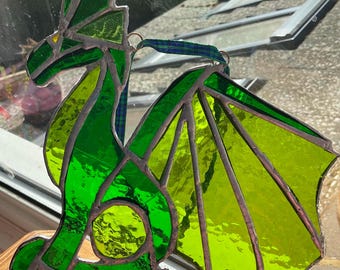 May include: A stained glass dragon in shades of green, with a vibrant lime green wing. The dragon has a detailed design with a dark outline and a blue and green plaid ribbon for hanging. The dragon is sitting on a wooden surface.