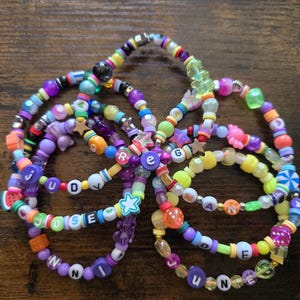 May include: A collection of colorful beaded bracelets in various sizes and designs. The bracelets feature a variety of beads in shades of purple, green, yellow, and orange, with letter beads spelling out names. The bracelets are arranged on a dark wooden surface.
