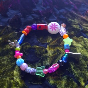 May include: A colorful beaded bracelet with a variety of shapes and colors. The bracelet includes red, pink, blue, and green beads, along with a white bead with a pink flower design and a silver skull charm.