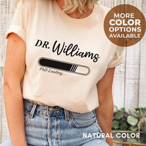 May include: A natural-colored t-shirt with the text "DR. Williams" above a loading bar graphic that reads "PhD Loading...". A circular graphic indicates more color options are available. The shirt is paired with blue jeans.