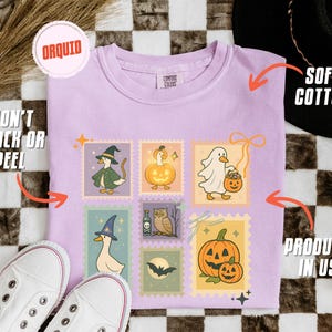 May include: A light purple t-shirt with a Halloween-themed stamp design featuring cartoon ducks in costumes and pumpkins. The shirt is made of soft cotton and is produced in the USA. The design won't crack or peel.