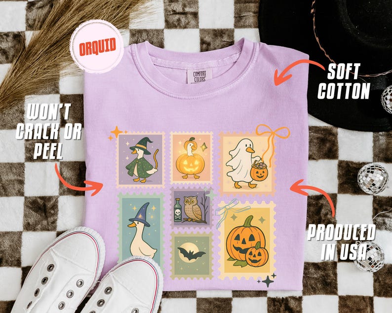 May include: A light purple t-shirt with a Halloween-themed stamp design featuring cartoon ducks in costumes and pumpkins. The shirt is made of soft cotton and is produced in the USA. The design won't crack or peel.