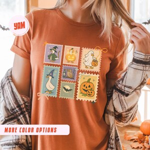 May include: Rust-colored t-shirt featuring a colorful Halloween-themed stamp design. The stamps depict various spooky characters like a witch, ghost, and pumpkin. The shirt also has the text "MORE COLOR OPTIONS" on the bottom.
