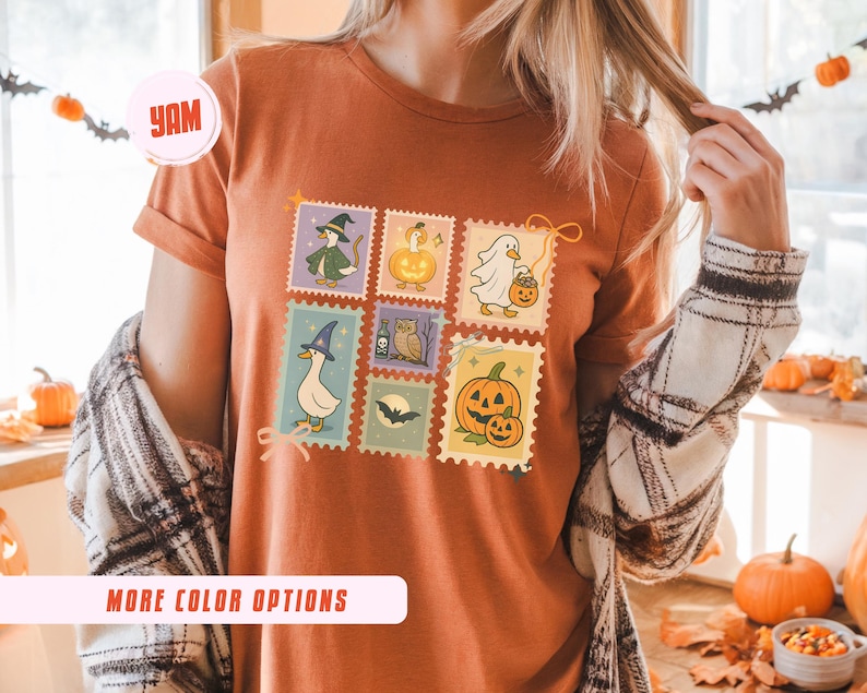 May include: Rust-colored t-shirt featuring a colorful Halloween-themed stamp design. The stamps depict various spooky characters like a witch, ghost, and pumpkin. The shirt also has the text "MORE COLOR OPTIONS" on the bottom.