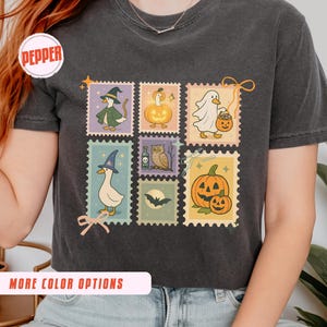 May include: Dark gray t-shirt featuring six postage stamp designs with Halloween-themed illustrations. The stamps depict a witch duck, a pumpkin, a ghost, a duck with a witch hat, an owl, and a pumpkin with a smaller pumpkin. The shirt has the word "Pepper" in a circle.