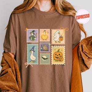 May include: Brown t-shirt featuring a collection of Halloween-themed postage stamp designs. The stamps depict a witch duck, a pumpkin, a ghost with a pumpkin bucket, a duck with a witch hat, an owl, a bat, and a jack-o'-lantern.