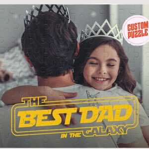 May include: A father and daughter are hugging. The daughter is wearing a silver crown and is smiling. The father is wearing a grey shirt. The text "THE BEST DAD IN THE GALAXY" is in yellow and white on a black background.