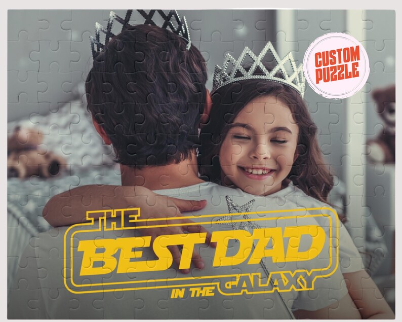 May include: A father and daughter are hugging. The daughter is wearing a silver crown and is smiling. The father is wearing a grey shirt. The text "THE BEST DAD IN THE GALAXY" is in yellow and white on a black background.