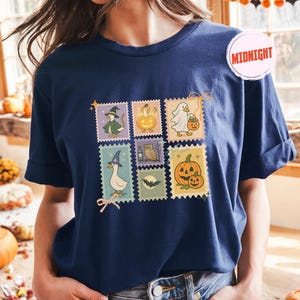 May include: Navy blue t-shirt featuring a colorful Halloween-themed stamp design. The stamps depict various spooky characters, including a witch, a ghost, and pumpkins. The word "Midnight" is printed on a pink circle on the shirt.