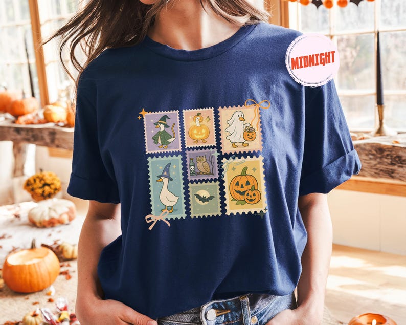 May include: Navy blue t-shirt featuring a colorful Halloween-themed stamp design. The stamps depict various spooky characters, including a witch, a ghost, and pumpkins. The word "Midnight" is printed on a pink circle on the shirt.