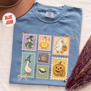 May include: A blue jean colored t-shirt featuring a collection of Halloween-themed postage stamp designs. The stamps depict cartoon ducks dressed as a witch, a ghost, and other Halloween characters. The shirt has a crew neck and short sleeves.
