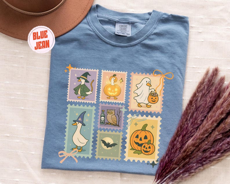 May include: A blue jean colored t-shirt featuring a collection of Halloween-themed postage stamp designs. The stamps depict cartoon ducks dressed as a witch, a ghost, and other Halloween characters. The shirt has a crew neck and short sleeves.