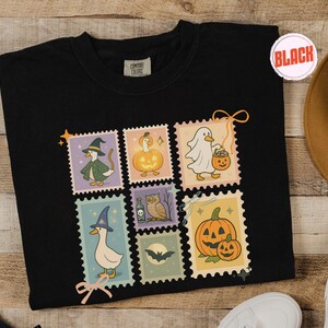 May include: Black Comfort Colors t-shirt featuring a collection of Halloween-themed postage stamp designs. Each stamp showcases a different whimsical illustration, including a witch duck, a ghost duck, and pumpkins. The shirt has a round neckline.