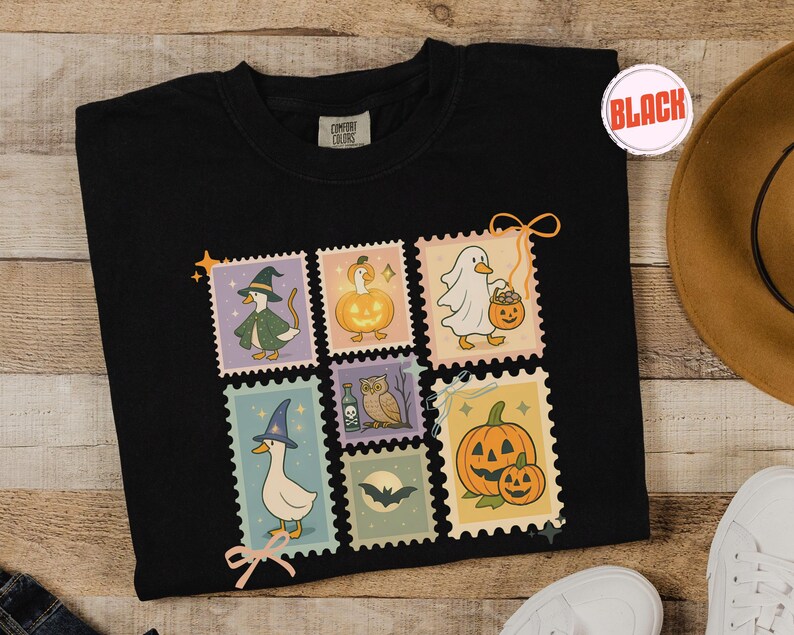 May include: Black Comfort Colors t-shirt featuring a collection of Halloween-themed postage stamp designs. Each stamp showcases a different whimsical illustration, including a witch duck, a ghost duck, and pumpkins. The shirt has a round neckline.