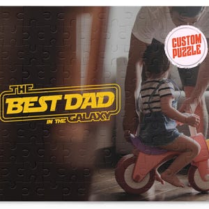 May include: A black and white jigsaw puzzle with a yellow and orange graphic that says "The Best Dad in the Galaxy". A man and a child are in the background, with the man holding a sign that says "Custom Puzzle".