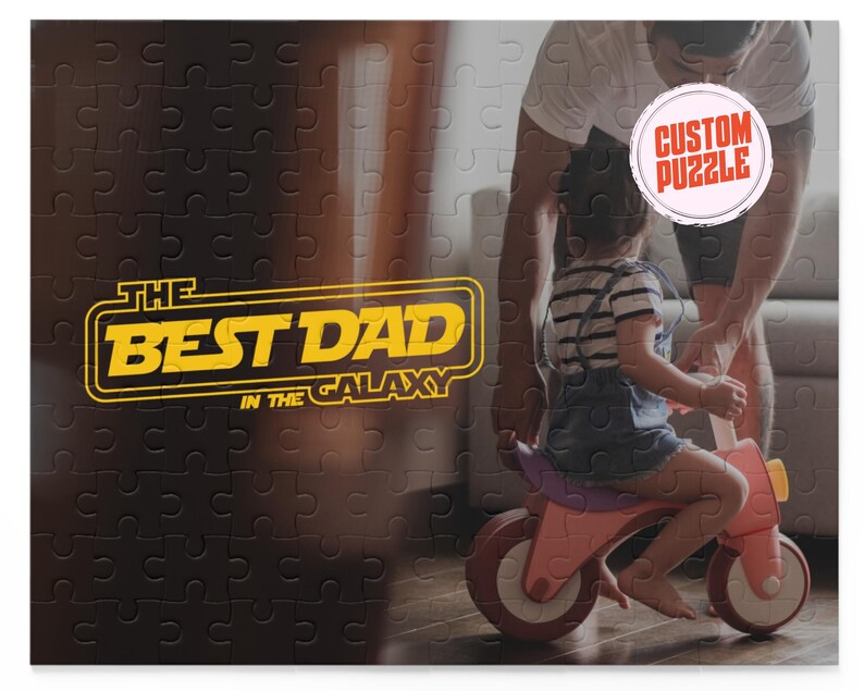 May include: A black and white jigsaw puzzle with a yellow and orange graphic that says "The Best Dad in the Galaxy". A man and a child are in the background, with the man holding a sign that says "Custom Puzzle".
