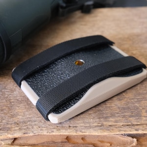 May include: A black and tan camera mount with two black straps. The mount has a textured surface and a small hole in the center.