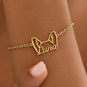May include: A gold-toned metal bracelet featuring a delicate chain and a central pendant shaped like the outline of animal ears with the name Luna written in a cursive script integrated into the design of the ears.