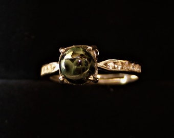 Op de afbeelding: Een gouden ring met een grote, ronde, groene edelsteen in het midden. De band is ook goudkleurig en bezet met kleine, heldere edelstenen. De ring staat tegen een donkere achtergrond, waardoor de details opvallen.