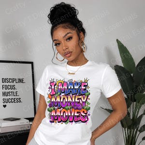 May include: White t-shirt with the phrase "I Make Money Moves" in a colorful graffiti-style design. The shirt features dollar bills and crowns. A framed print with the words "Discipline, Focus, Hustle, Success" is visible in the background.