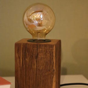 Puede incluir: Lámpara de mesa rústica de madera con una gran bombilla Edison redonda de color ámbar. La bombilla se asienta sobre una base cuadrada de madera maciza. La lámpara tiene un cable de alimentación negro. La base de madera mide unos 10 cm de ancho.