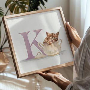 Op de afbeelding: Ingelijste aquarelprint van een slapend katje in een theekopje, met de letter "K" in lavendel. Het kunstwerk is in een licht houten frame. Het katje heeft bruine en beige strepen.