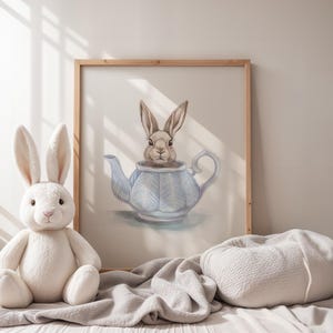 Puede incluir: Una ilustración de acuarela de un conejo marrón sentado dentro de una tetera azul y blanca. La ilustración está enmarcada y colgada en una pared sobre una cama blanca con una manta gris y una almohada gris.