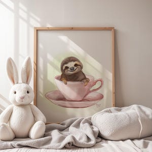 May include: A white plush bunny rabbit sits in front of a framed print of a sloth in a pink teacup. The print has a light green background and the text "Nina Lee" in the bottom right corner.