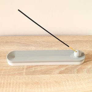 May include: A grey and white ceramic incense holder with a round, grey top. A black incense stick is resting on the top of the holder.