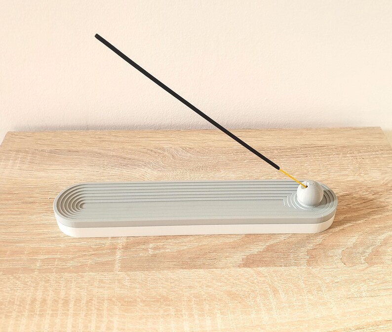 May include: A grey and white ceramic incense holder with a round, grey top. A black incense stick is resting on the top of the holder.