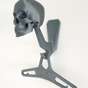 May include: A gray 3D-printed skull-shaped wall hook with a hanger design. The skull is mounted on a bracket, and the hanger is attached below. The hook is designed to hold clothing or accessories. The design is modern and minimalist.