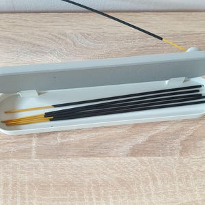 May include: A white rectangular incense holder with a lid, open to reveal a set of black incense sticks with gold tips.