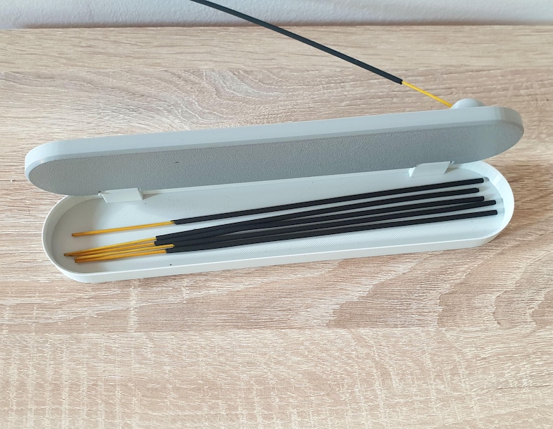 May include: A white rectangular incense holder with a lid, open to reveal a set of black incense sticks with gold tips.