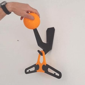 May include: A black wall-mounted hook with an orange hanger attached. A hand is holding an orange ball above the hook.