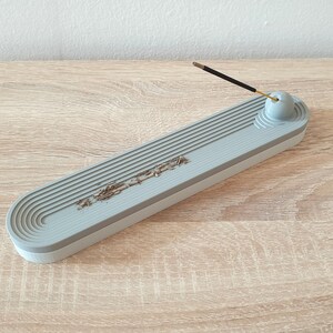 May include: A grey ceramic incense holder with a grooved design and a round knob on the end. The holder is designed to catch ash from incense sticks.